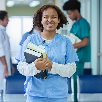 A nurse student in scrubs smiling and holding books in her hands.