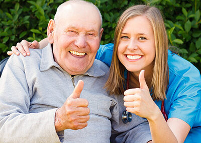 A senior in a wheelchair holding a thumbs up with a nurse beside him.