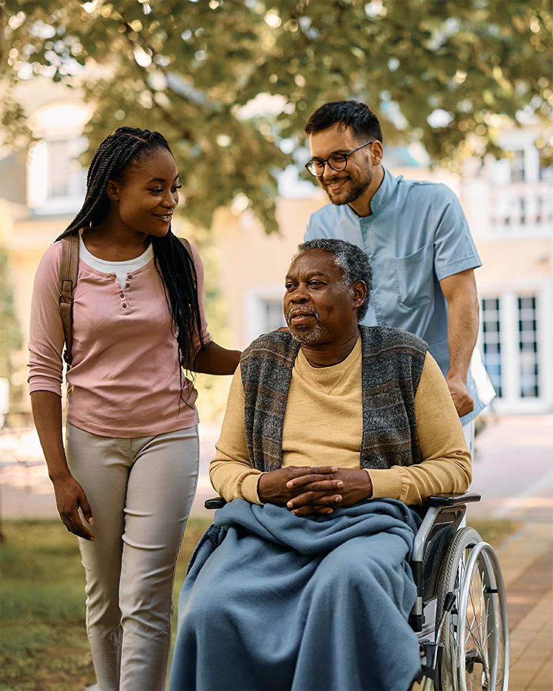 A mature male and his children walking him outside in his wheelchair.