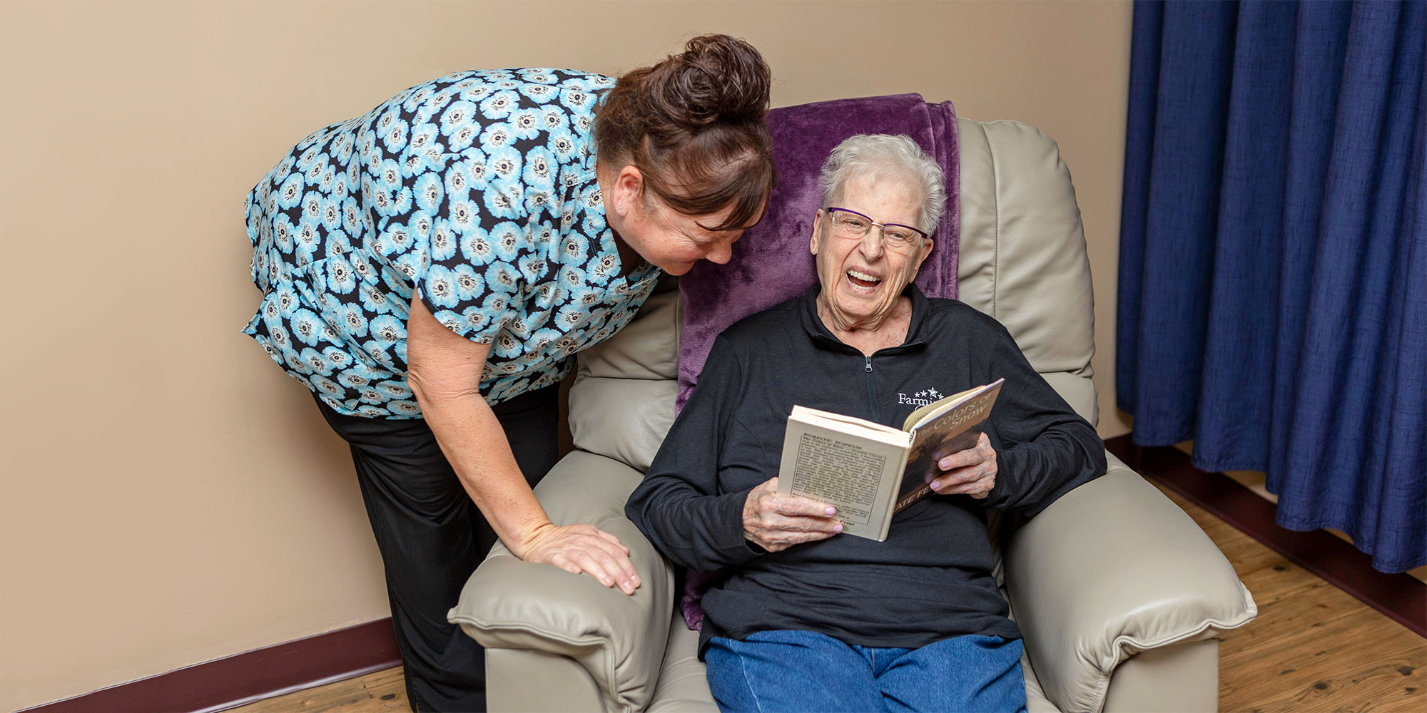 Woman reading a book and a caregiver smiling and laughing together at the Farmington Village facility